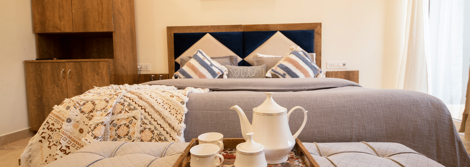 A hotel room, featuring a large grey bed with white linens, blue and orange pillows, a wooden tray with a teapot and cups on the bed, and a painting on the wall | Perfectstayz Value Hills, Rishikesh