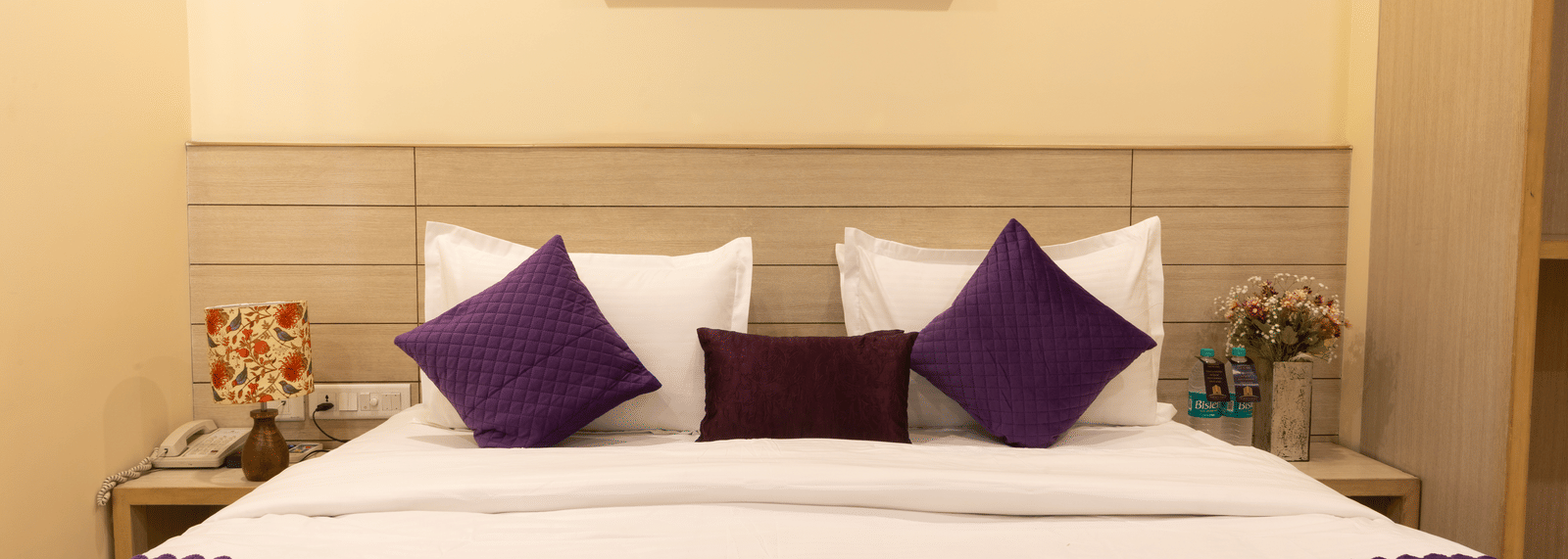 A close-up of a large bed, featuring a white duvet, purple runner and pillows, a light wooden headboard, and a small framed painting on the wall | Perfectstayz Value Sagar