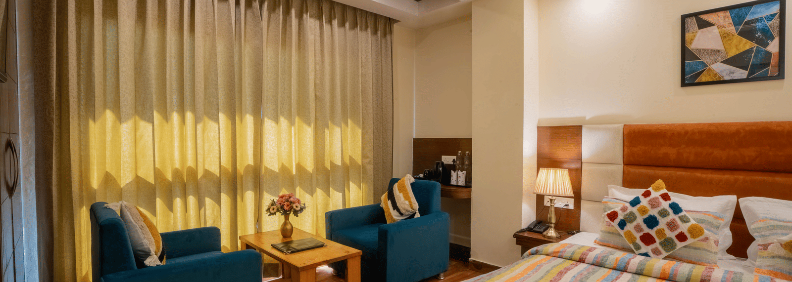 A hotel room, featuring a large bed with a striped duvet, thick beige curtains, and 2 teal blue chairs with a small table | Perfectstayz Value Valley Vista