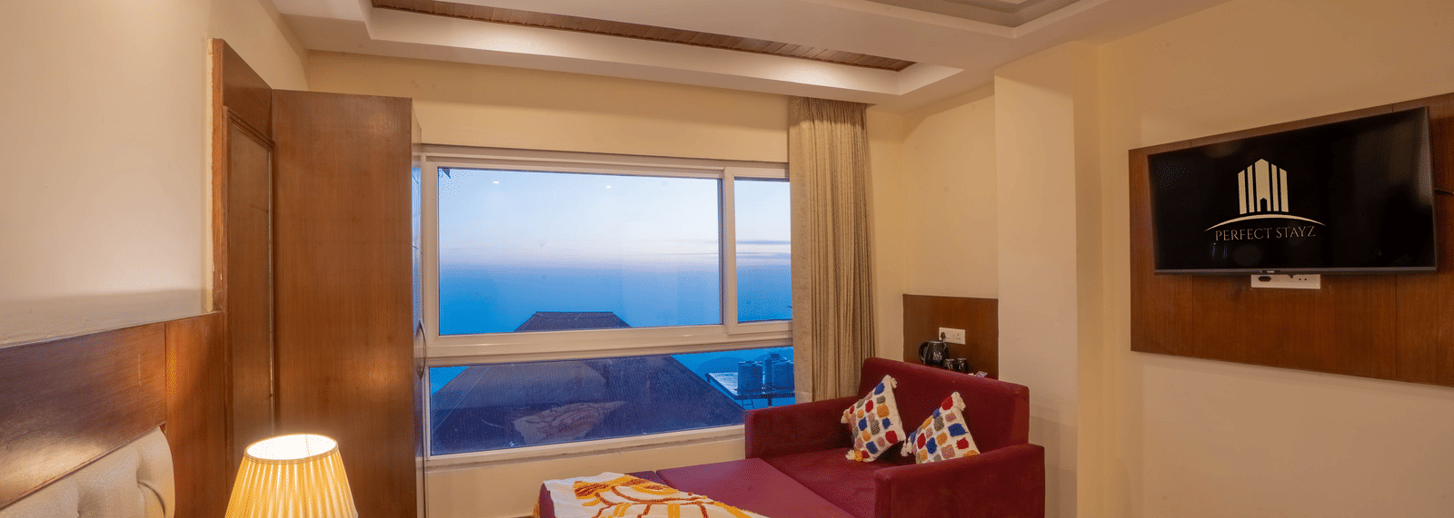 A hotel room with a large window view of a hilly landscape, featuring a bed with white linens, a wall-mounted TV, and a red sofa | Perfectstayz Value Valley Vista