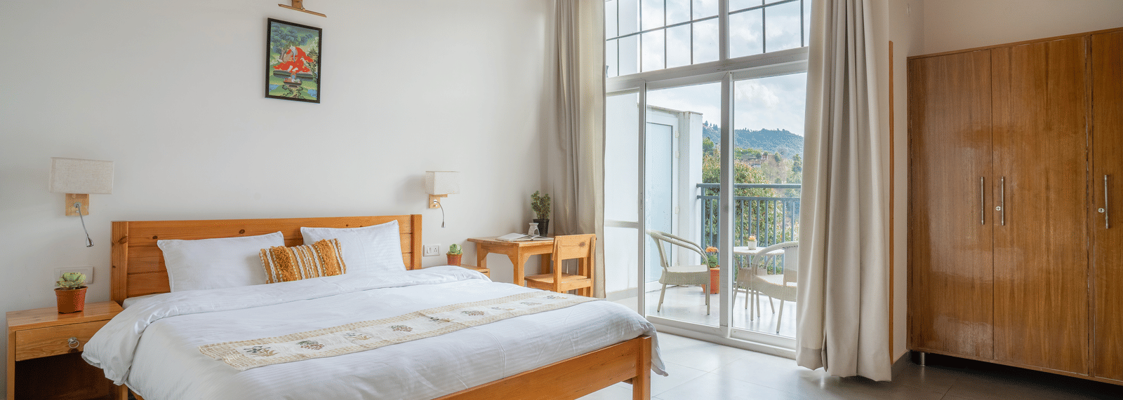 A wide shot of a Ziran Retreat bedroom with a large bed, a wooden bedside table, and large glass doors open to a balcony with a scenic view.