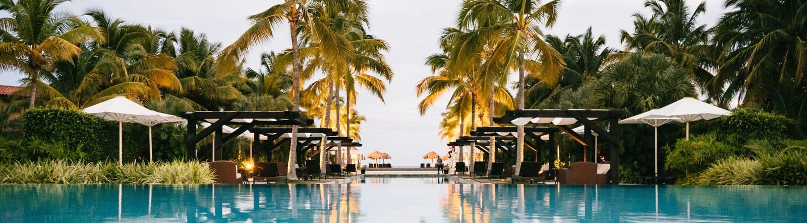 Expansive resort swimming pool reflecting the sky and surrounding palm trees and cabanas.