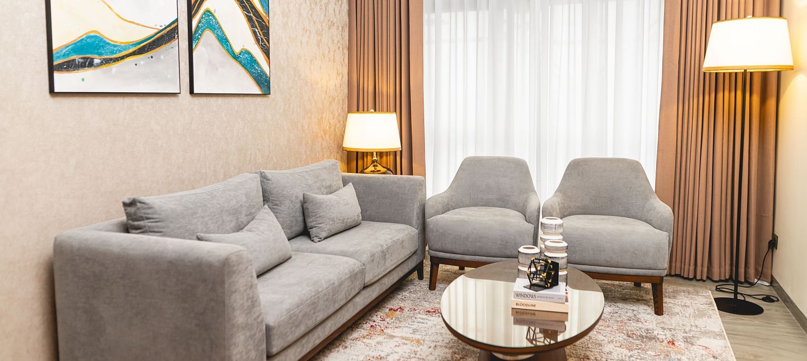 Room at KertelSuites with a sofa, two matching armchairs, a round coffee table, two floor lamps, a patterned rug, and abstract art on the wall. Large windows with sheer curtains allow natural light in.