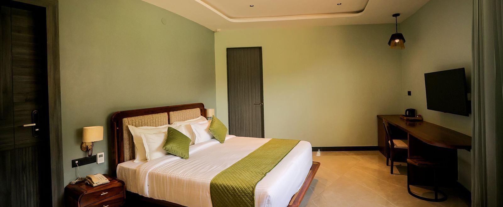 Valley View Room furnished with a king size bed dressed in white linens and a green accent runner, complemented by a wooden side table and a television mounted on the wall at The Qualia Resort Club & Brewery, Udaipur.