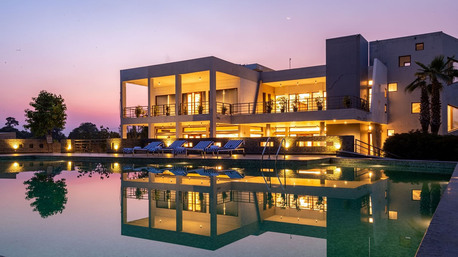 Facade view of DLS Forest Vines Resort, with warm light glowing from its windows and doors, reflected on the shimmering swimming pool beneath a clear sky.