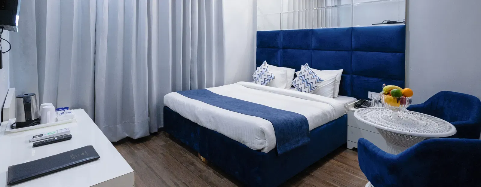 Deluxe Room featuring a king size bed with a blue headboard and white and blue linens, two blue chairs with a small round table, and light wood-look flooring. - Hotel Downtown17, Chandigarh.