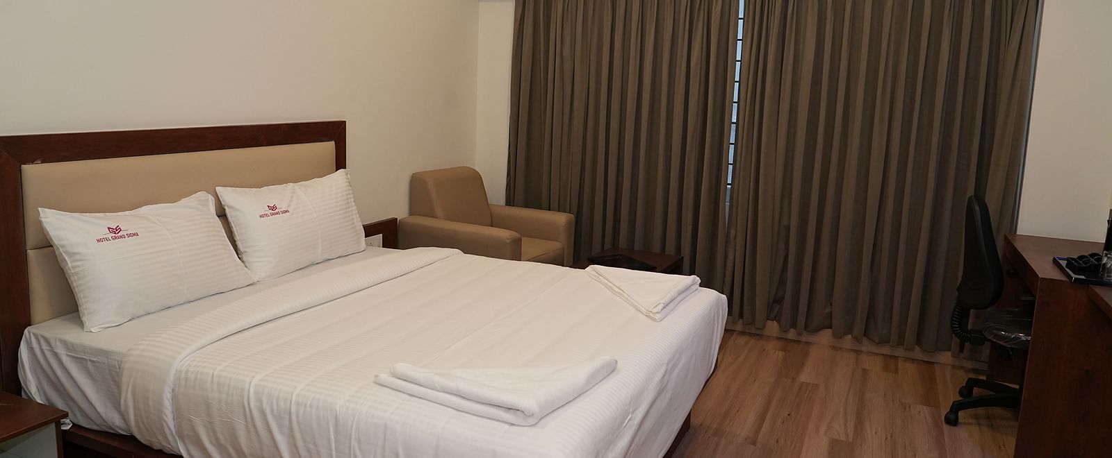 corner view image of the double bed with a huge window in the corner covered with brown curtains - Hotel Grand Sigma