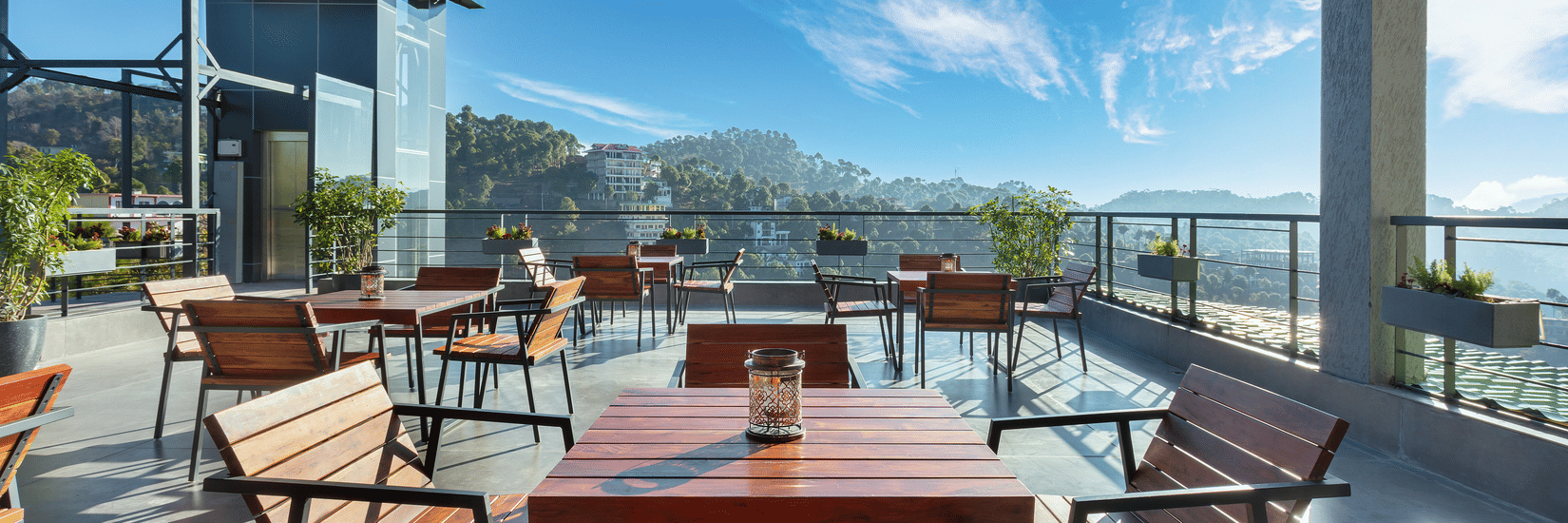 Outdoor dining area at Suryavilas Luxury Resort and Spa, featuring wooden tables and chairs on a spacious terrace with panoramic views of the Himalayan hills under a bright blue sky.