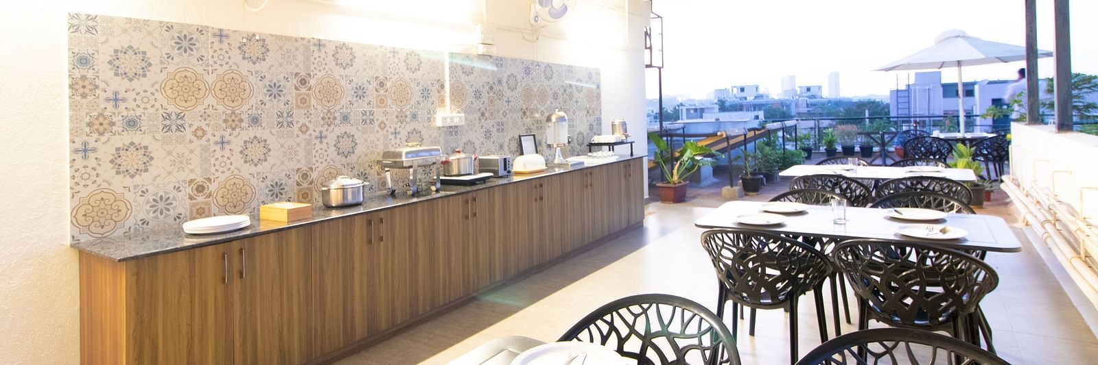 A dining space with tables and chairs under a partial roof, featuring a buffet setup along one side at Sanctum Suites Indiranagar, Bangalore.