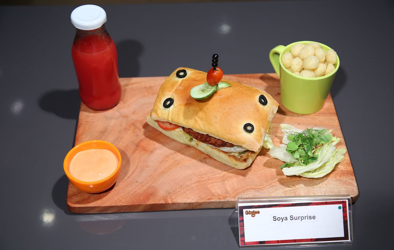  A wooden board holding a sandwich, a small cup of potato wedges, a bottle of juice, and dipping sauces.