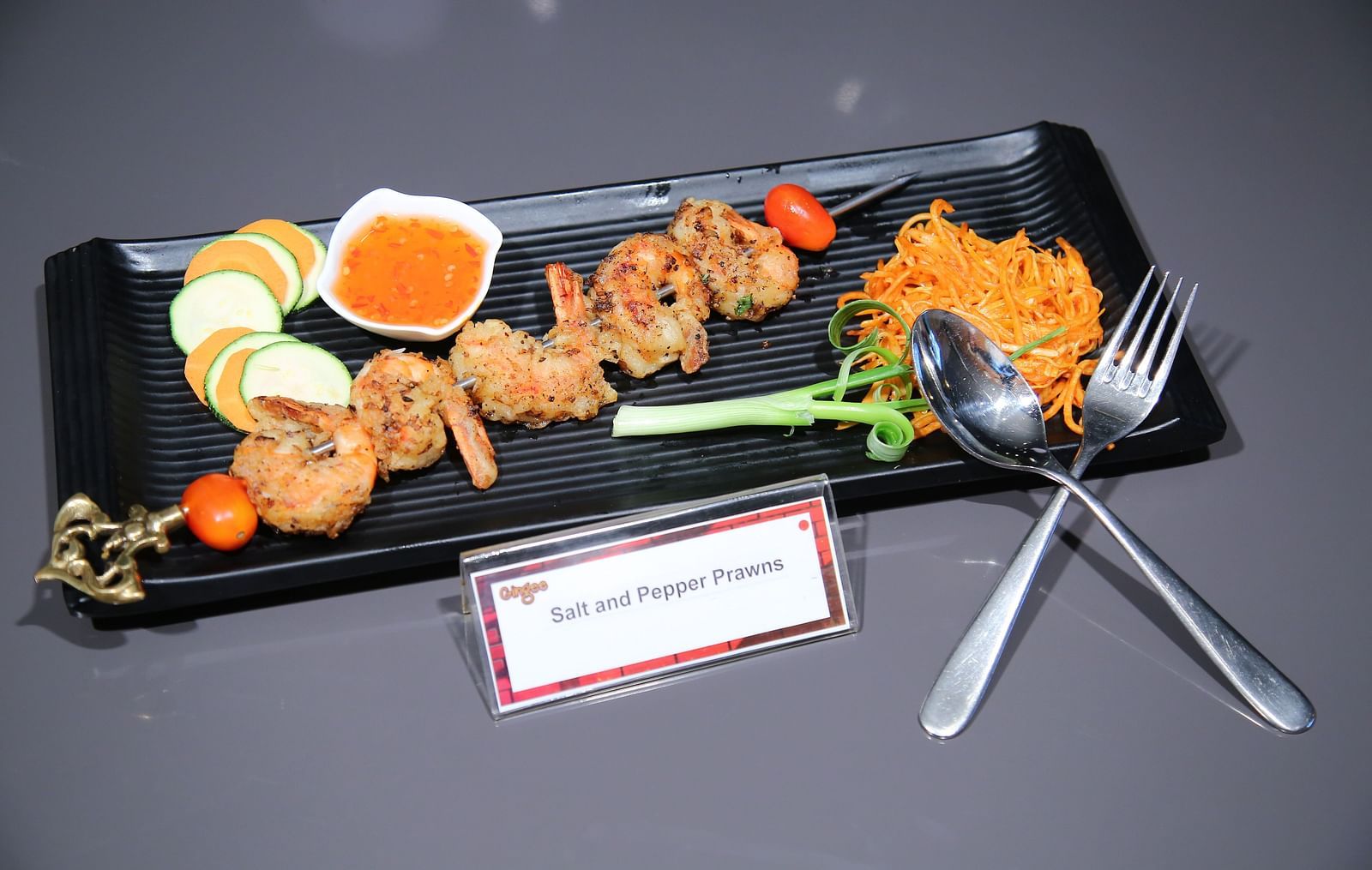A black rectangular plate with three food items. On the left, there are stir-fried vegetables and what appears to be meat skewers. In the center, a square dish with a golden-brown topping. On the right, a small bowl of red sauce.