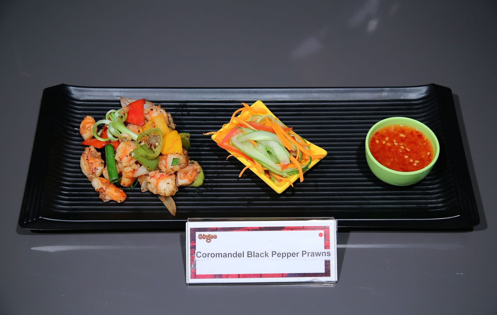 A black rectangular plate with three food items. On the left, there are stir-fried vegetables and what appears to be meat skewers. In the center, a square dish with a golden-brown topping. On the right, a small bowl of red sauce.