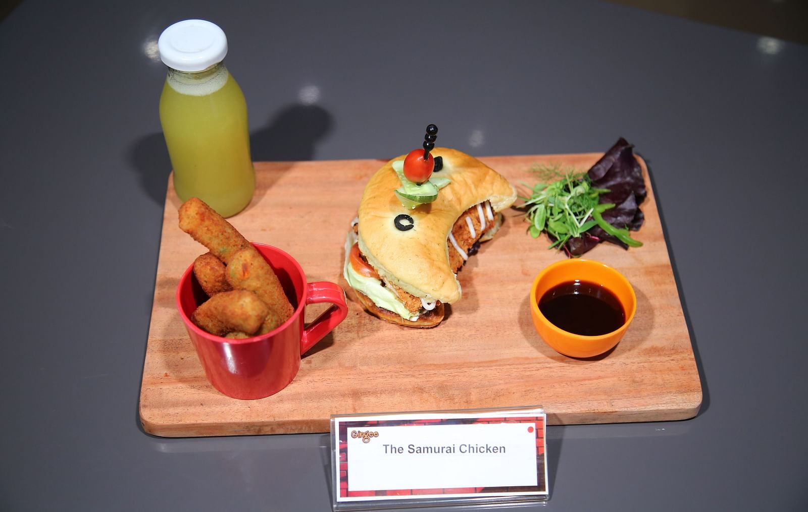 A wooden board with a sandwich, crispy chicken tenders in a red cup, a dipping sauce, and a bottle of juice.