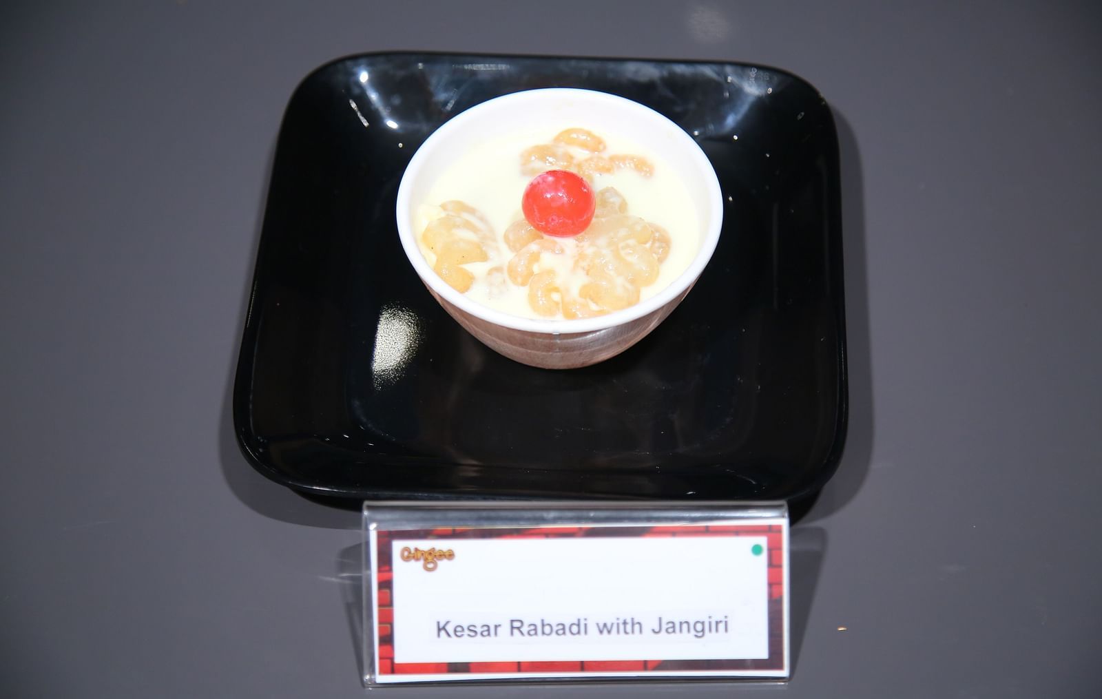 A white bowl filled with a creamy dessert topped with a red cherry on a black square plate.