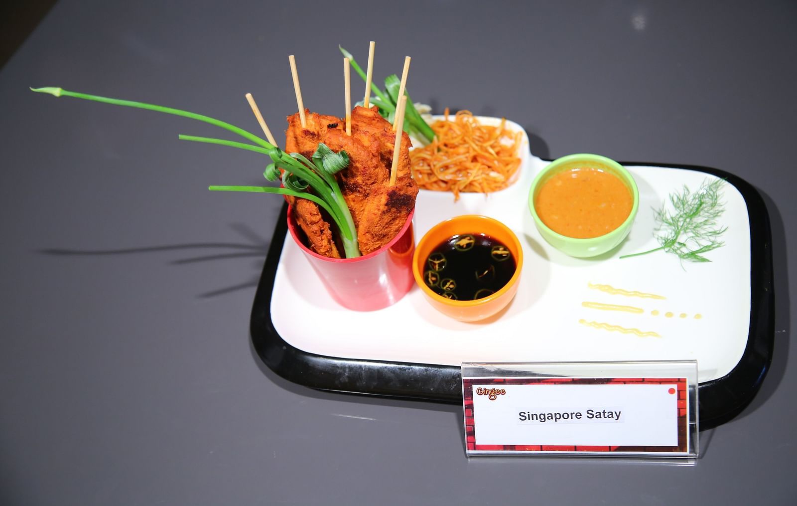 A white rectangular plate with three red cylindrical cups containing skewers of what appears to be meat, served with two dipping sauces in small bowls. The skewers are garnished with green stalks.
