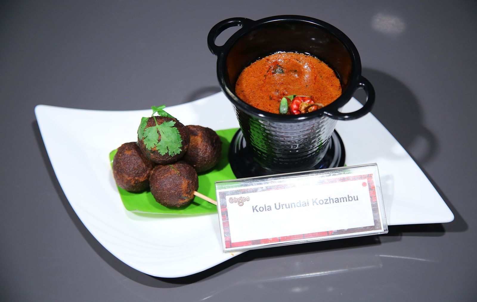A white plate with round, dark brown appetizers on skewers, served with a small, red, circular bowl of dipping sauce.