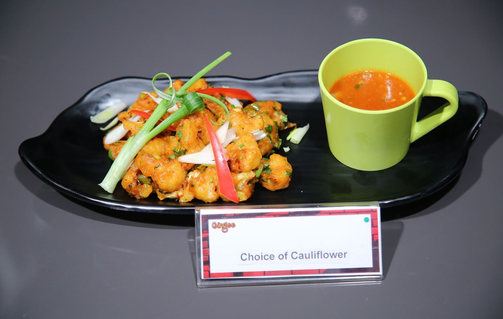 A black oval plate with a mound of crispy fried vegetables, garnished with green onions and red peppers. A small green cup of soup is placed beside it. A label at the bottom reads Crispy and Crunchy.