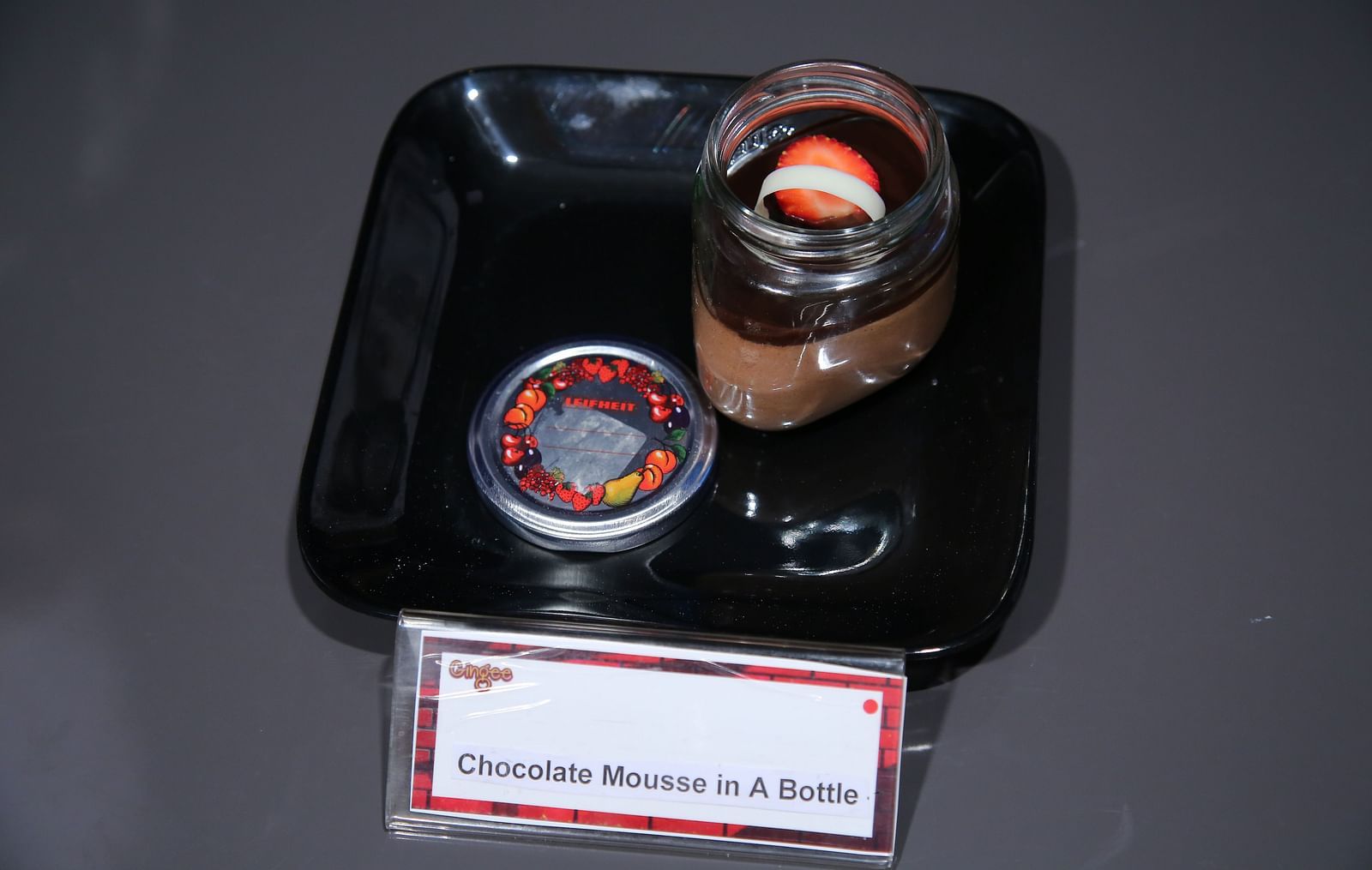 A glass jar filled with chocolate mousse, topped with a small, round object. A black tray holds the jar and a circular coaster.