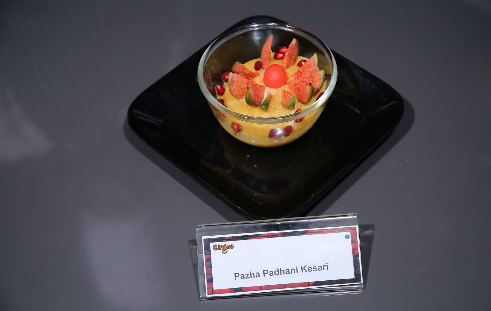  A clear glass bowl filled with a yellow custard-like dessert, topped with a cluster of red berries. The dish is placed on a black square plate with a label reading Passion Fruit Pudding Crispy.