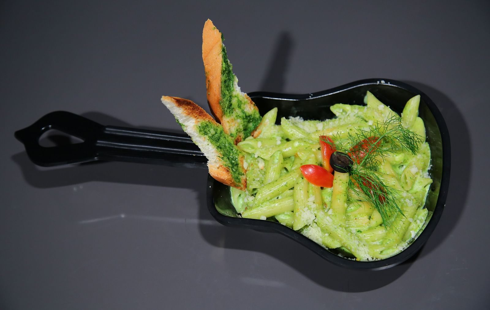 A black guitar-shaped plate filled with a creamy green pasta dish. The plate has a handle at one end and a guitar head at the other. The pasta is topped with a red chili. 