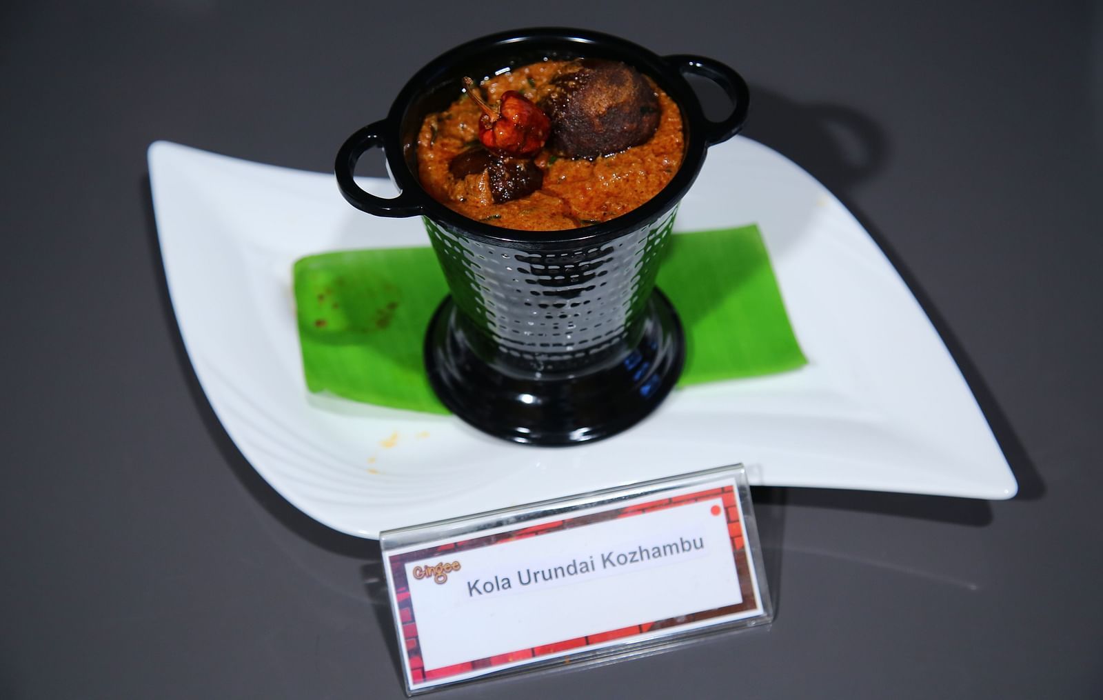 A bowl of stew with a red pepper and a cinnamon stick, served on a white plate with a green leaf.
