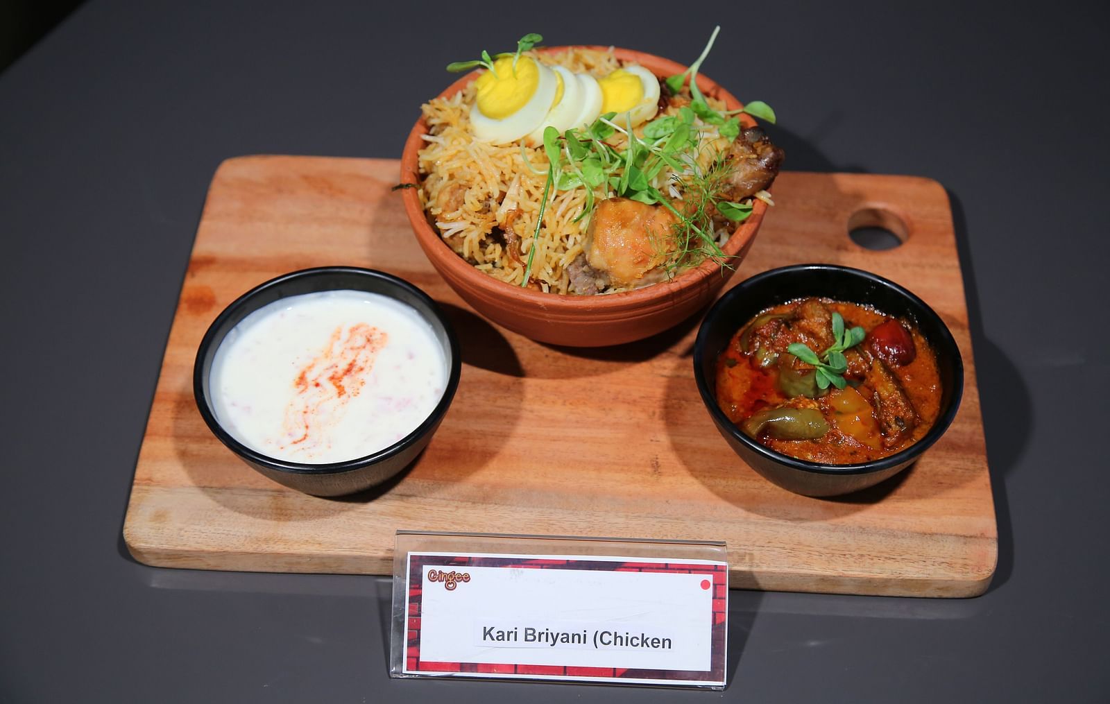 A wooden platter with three bowls. The center bowl contains noodles, half a boiled egg, and garnishes. The left bowl holds a white creamy liquid, and the right bowl contains a reddish curry.