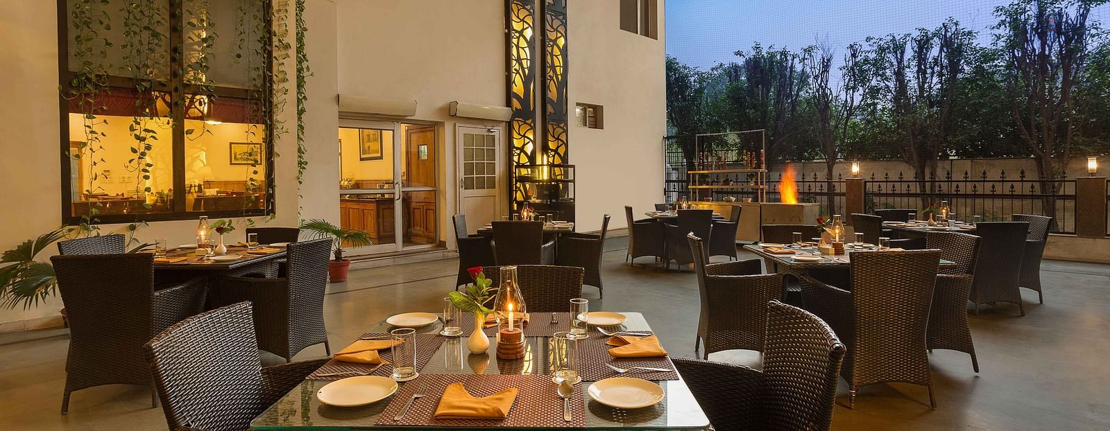 An open dining space on a terrace, featuring tables and chairs arranged with plates and cutlery, illuminated by soft lighting in the evening ambiance