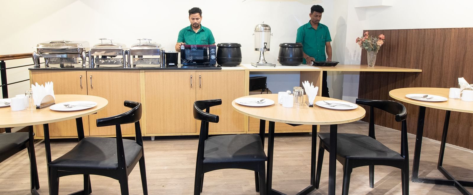 A breakfast buffet area with staff behind the counter, food warmers, and tables with black chairs - Sanctum Suites Richmond Road, Bangalore