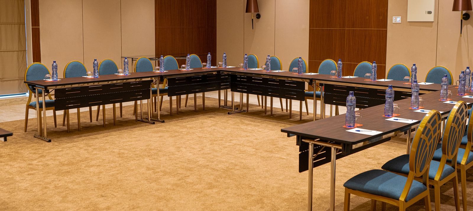 A conference room at KertelSuites with a U-shaped arrangement of tables and chairs, each table set with bottled water and notepads, in a well-lit, carpeted space