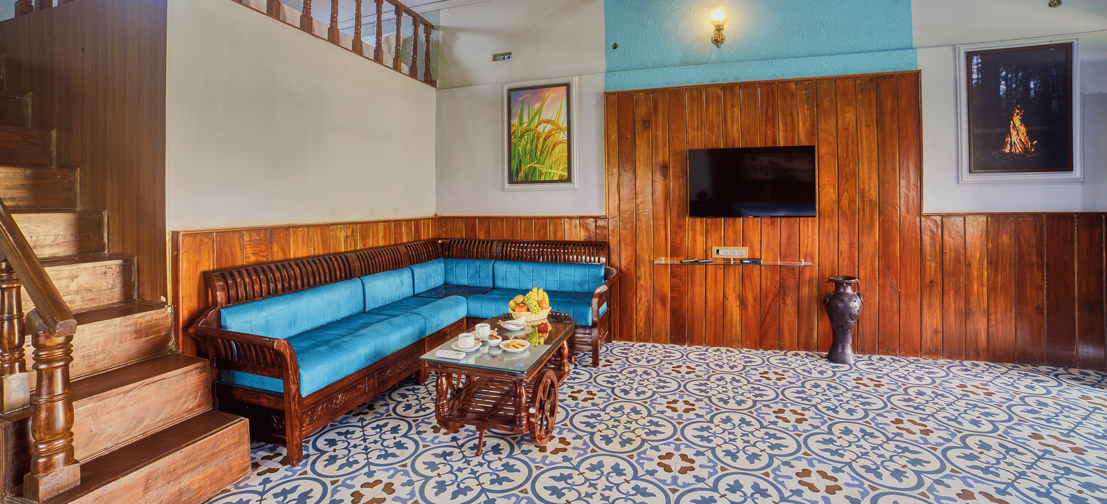 The interior of a spacious living area with patterned tiled floor, featuring a blue curved sofa, a wooden TV stand, and stairs leading to an upper level.
