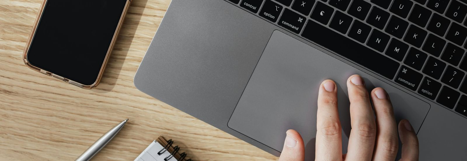 A person's hand is on the track pad of a laptop, with a smartphone and a notebook visible on a wooden desk.