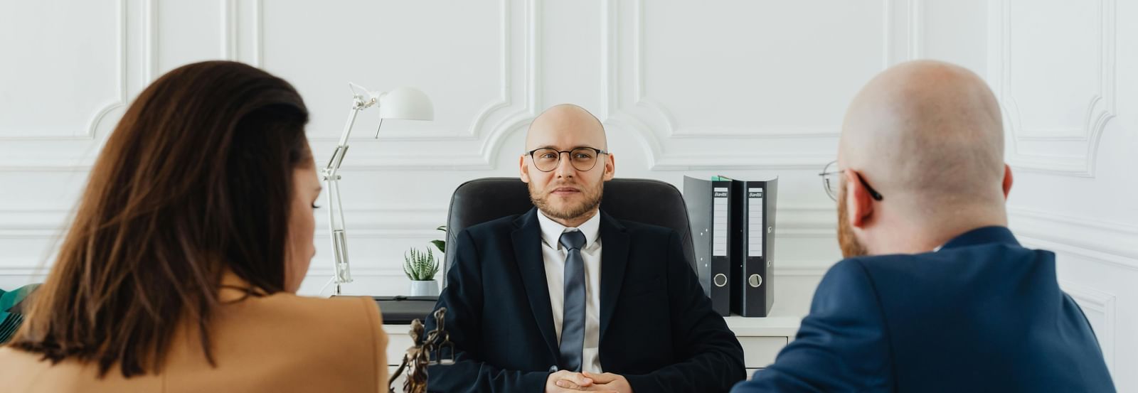 A person in a suit and eyeglasses sits at a desk, facing two other individuals who are seated opposite to him in a professional office setting. 