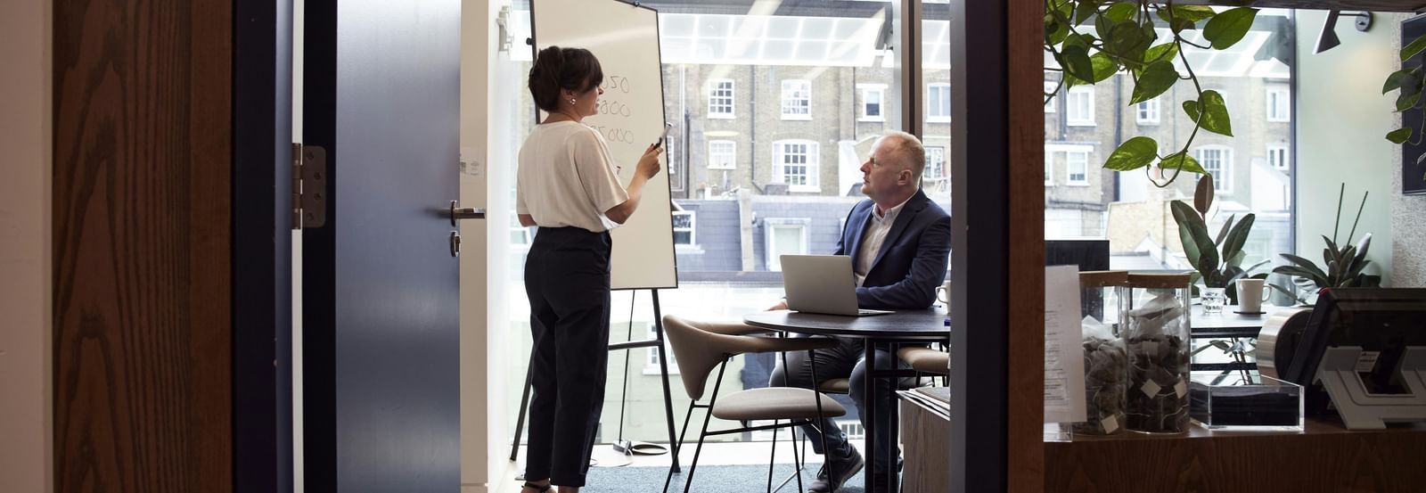 A person presents on a whiteboard to another person seated at a desk in a well-lit office setting.