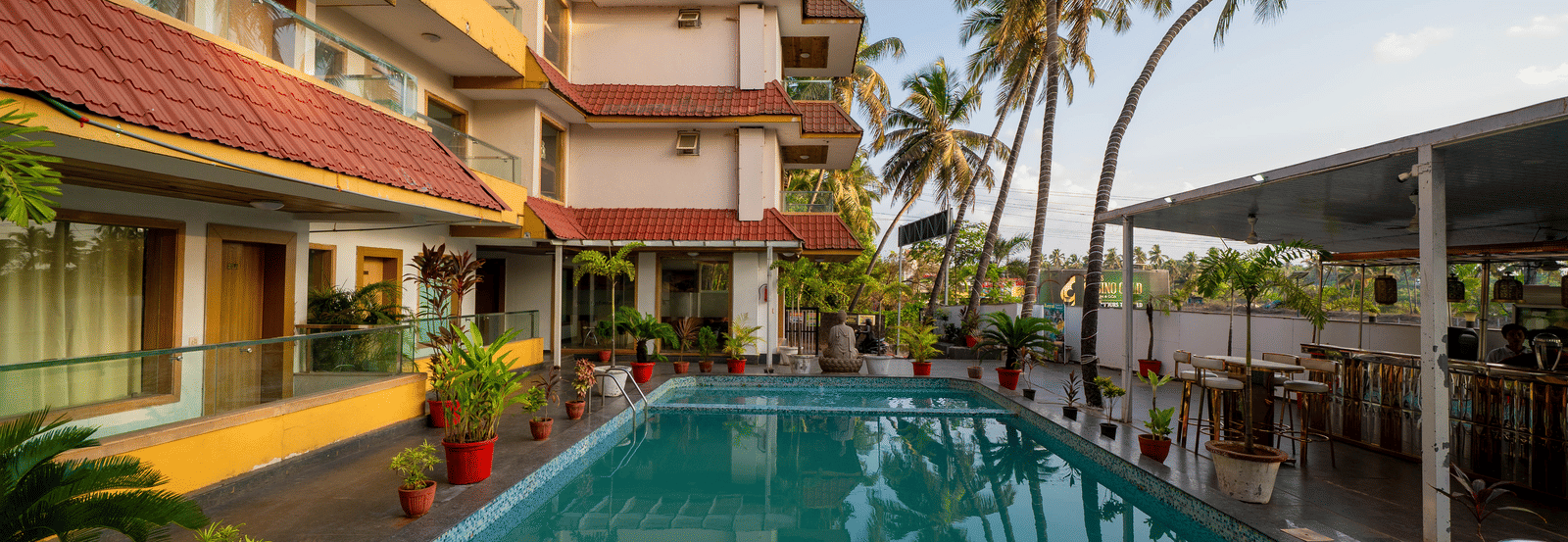 A sunny outdoor swimming pool is located within the courtyard surrounded by palm trees and buildings at Perfectstayz Premium Goa Continental.