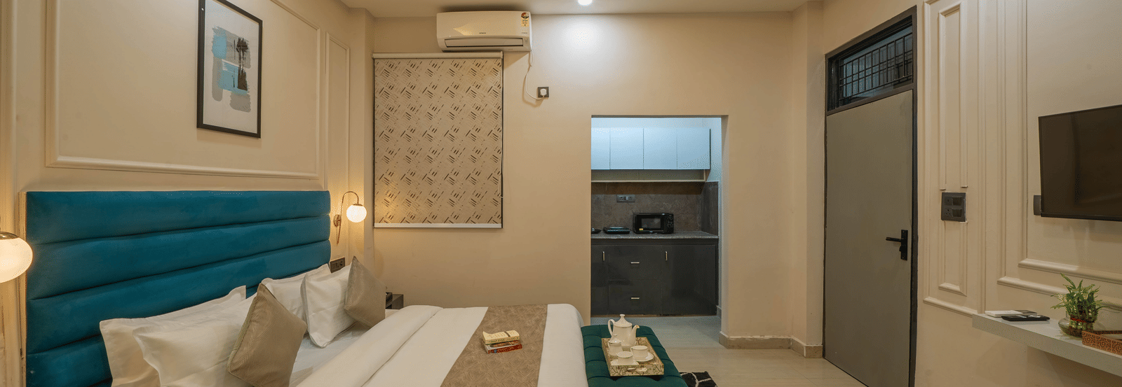 Spacious room with a bed, a teal tufted headboard, a white wardrobe, and a small kitchenette with dark grey lower cabinets next to the sleeping area - Perfectstayz Value Aalaya Suites, Haridwar