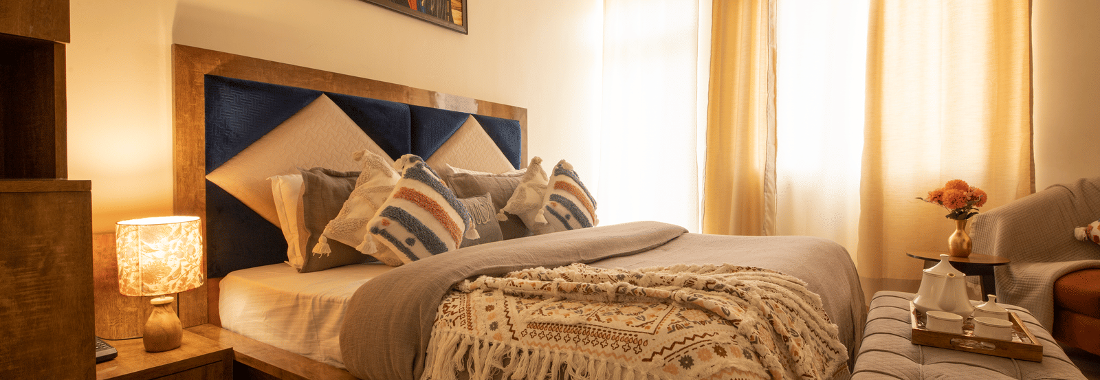 A brightly lit hotel room, featuring a large bed with a wooden frame, multiple decorative pillows, a knit throw blanket, and a window with sheer curtains | Perfectstayz Value Hills