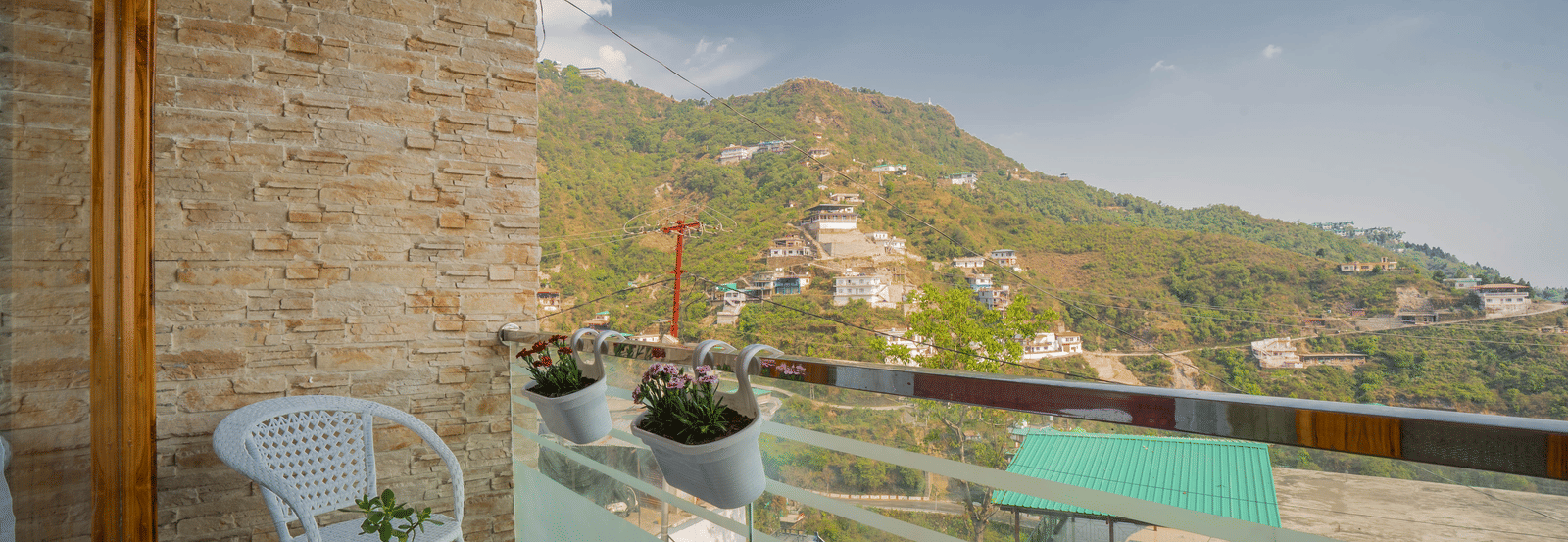 A balcony at Perfectstayz Value Paradise, Mussoorie, featuring a white chair, small table, and potted plants overlooking scenic hillside views and rooftops.
