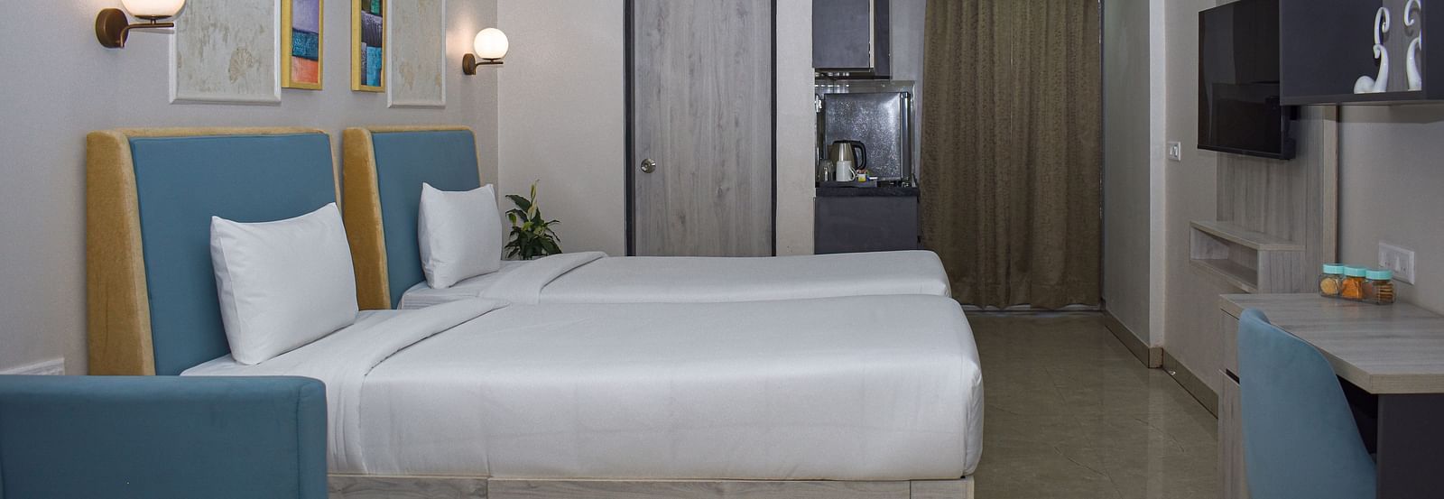 A hotel room with two twin beds, a blue sofa, a coffee table, a desk and chair, a wall-mounted TV, and a kitchenette area - Urban Suites by BluSalzz, Jaipur