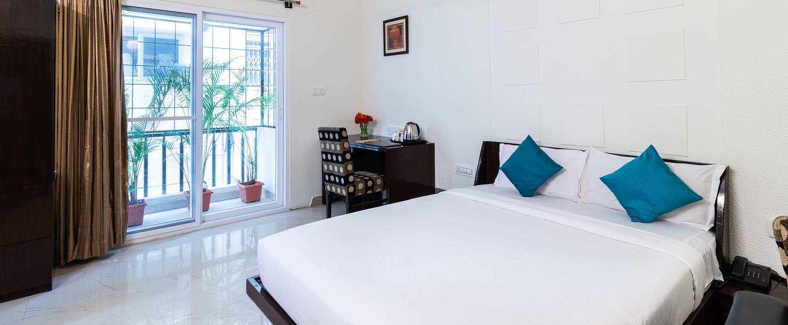 A room with a double bed with blue pillows, a work desk and chair, and glass door leading to the balcony at Sanctum Suites Domlur, Bangalore.