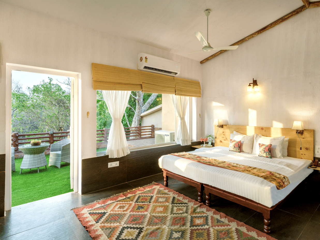 Premier Riverview Suite Twin at The Riverwood Forest Retreat, Pench featuring double size bed, bedside table with telephone, a window with blinds and a curtain, and a balcony with seating area.
