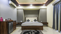 A modern hotel room with a double bed, padded headboard, side tables, mini-fridge unit with kettle, wall AC, ceiling lights, patterned rug, and floor-length curtains along the right side - Magnus Vosiv Suites, Kalyani Nagar