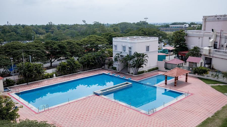A large outdoor swimming pool with clean water, surrounded by a tiled deck, nearby buildings, and trees under a cloudy sky at Jenneys Residency