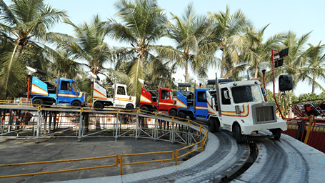 Gracy Bus ride with blue, white and red truck-shaped cars on a raised circular track, surrounded by yellow railings and palm trees at MGM Dizzee World, Chennai.