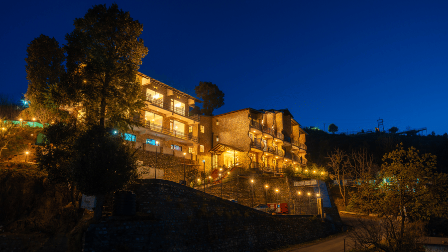 A wide shot of the multi-level resort building illuminated by warm lights against a dark blue night sky at Himalayas Resort By The Lake Hill, Mukteshwar.