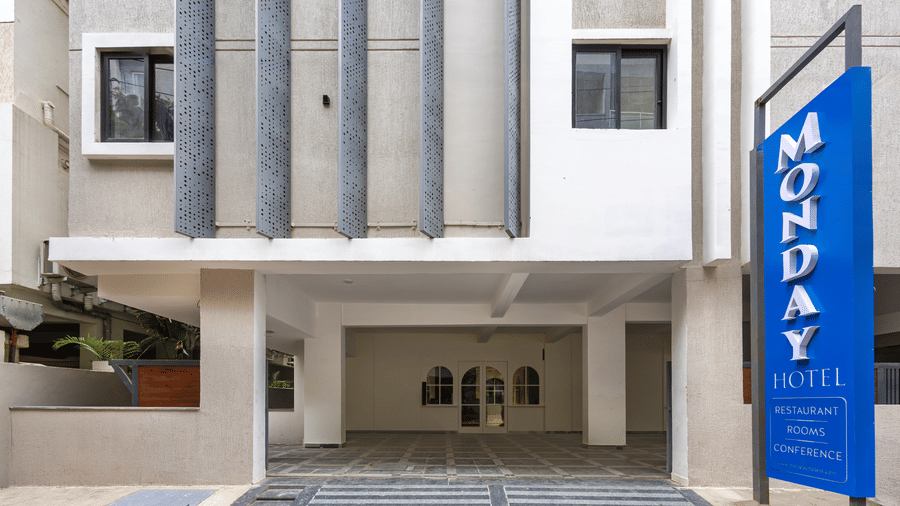 A building exterior of Monday Hotels & Suites Madhapur Hyderabad, featuring a blue sign reading 'MONDAY HOTELS' standing next to a covered ground entrance.