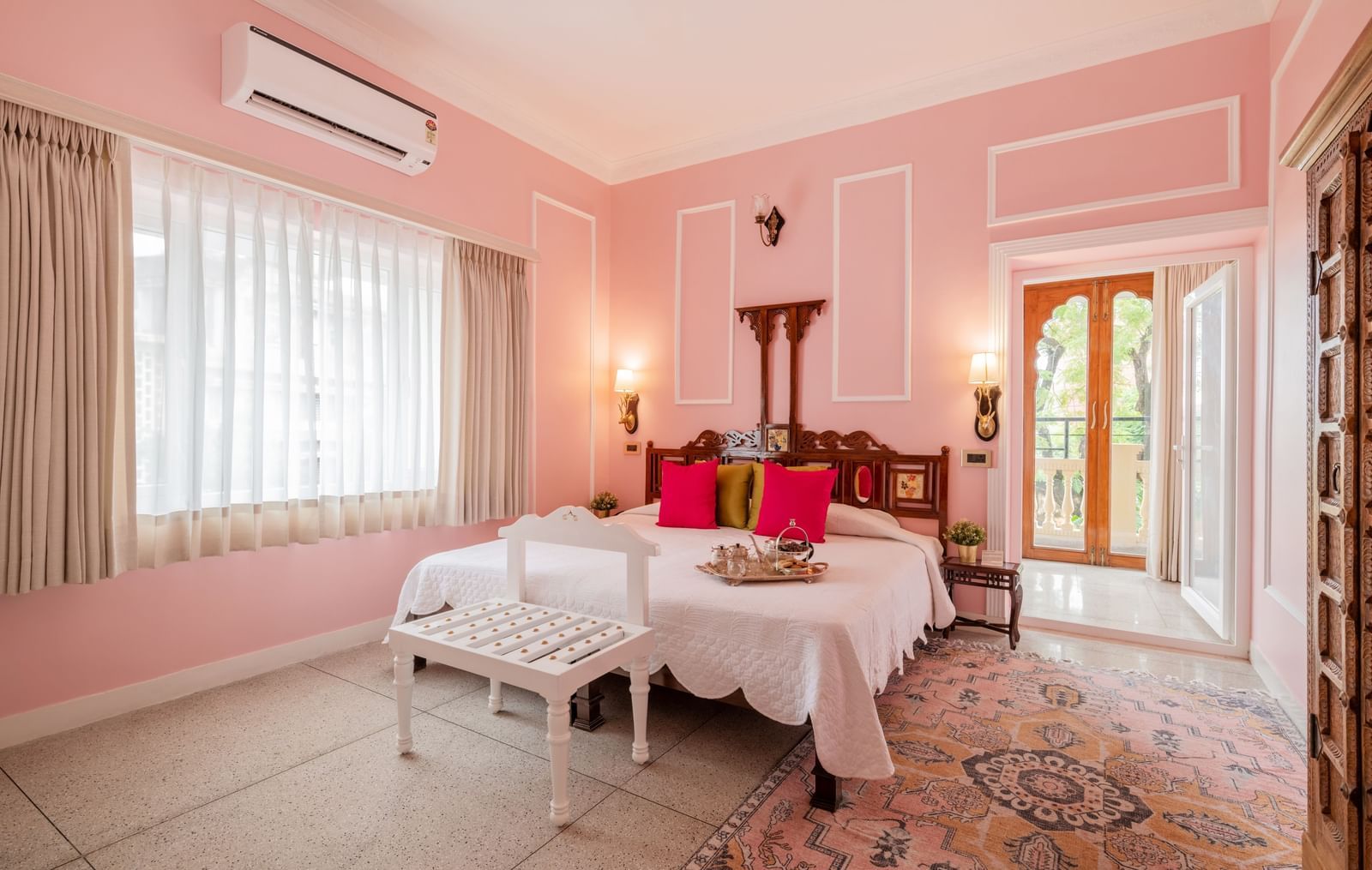 Pink-walled guest room with white bed, decorative cushions, and seating area at Dera Rawatsar, Jaipur.
