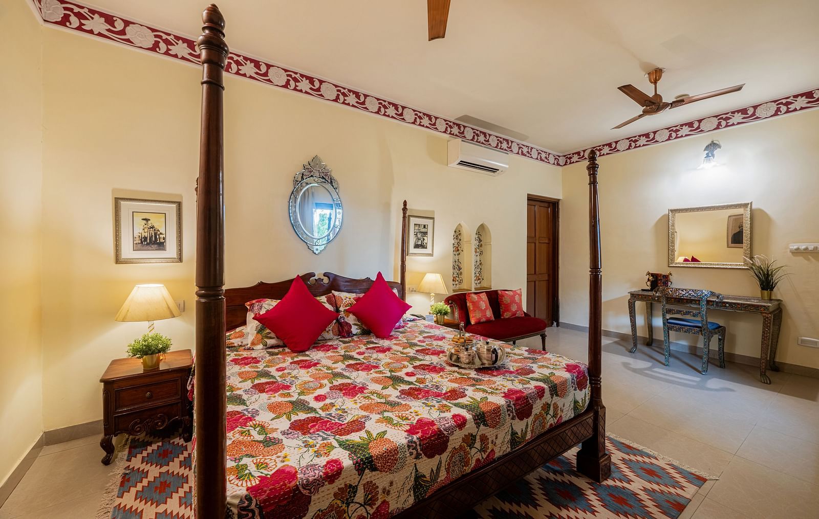 A comfortable bedroom at Dera Rawatsar in Jaipur includes a four-poster bed, a seating area, a dressing table, and decorative wall accents.