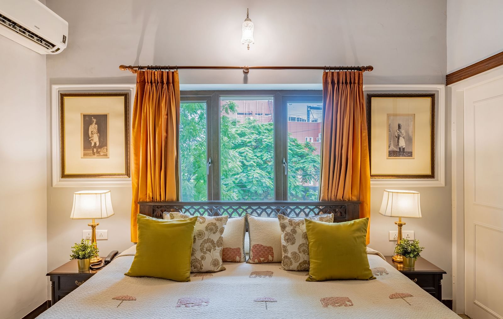 A tranquil bedroom at Dera Rawatsar in Jaipur features a comfortable bed, bedside lamps, framed artwork, and a window with vibrant curtains.