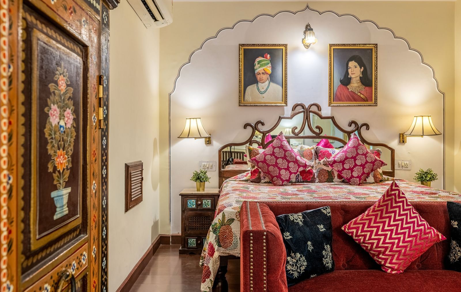 A beautifully decorated room at Dera Rawatsar, Jaipur, features a comfortable bed, a stylish loveseat, and an intricately painted wooden cabinet.
