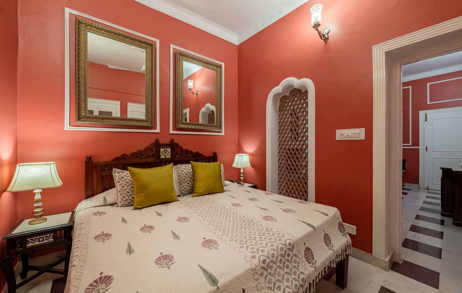 A vibrant bedroom at Dera Rawatsar, Jaipur, features a comfortable bed, framed mirrors, bedside lamps, and an ornate niche in the wall.